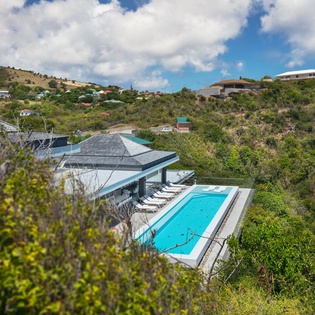 Modern hillside villa with pristine pool nestled among lush tropical vegetation and neighboring homes.