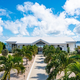 Contemporary beachfront villa welcomes guests along palm-lined pathway, framed by lush tropical landscaping and distant mountain views beneath vibrant Caribbean skies.