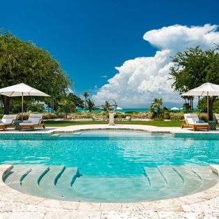 A stunning pool oasis with crystal-clear turquoise water, shaded loungers, and lush tropical surroundings framed by ocean views and dramatic Caribbean skies.