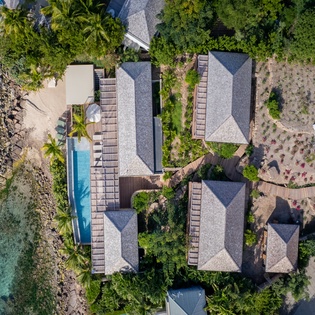 Stunning aerial view of beachfront resort buildings nestled among tropical palms, with crystal-clear turquoise waters and swimming pool.