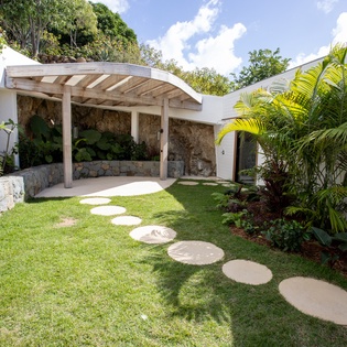 Tropical garden courtyard features stone pathways, lush palms, and a covered seating area beneath blue skies.