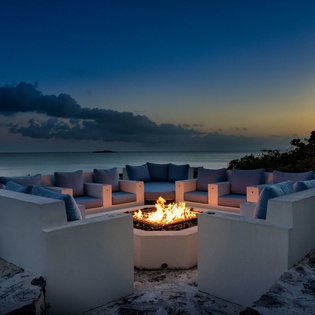 A stunning outdoor fire pit surrounded by built-in seating overlooks tranquil waters as twilight colors blend into night, providing an elegant beachside gathering spot.