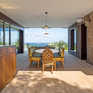 Dine in tropical elegance with ocean views through floor-to-ceiling windows, surrounded by natural stone and warm wood.