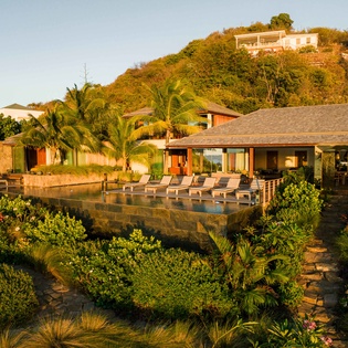A private tropical villa surrounded by lush landscaping, with a poolside terrace and hillside setting. The property offers secluded luxury in a natural, verdant environment.