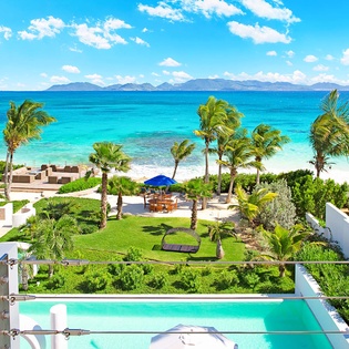 Beachfront paradise with pristine white sand, turquoise waters, and lush tropical gardens framed by distant mountain views under clear Caribbean skies.