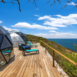 Stunning coastal property featuring geodesic dome structures perched on a hillside above the turquoise waters, with spacious wooden deck and lounge seating.