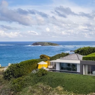 This stunning modern property sits on a coastal hillside overlooking the sparkling ocean, with a private island visible just offshore and lush native landscaping framing the sleek architecture.