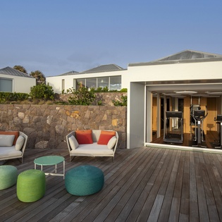 Modern outdoor fitness pavilion with exercise equipment opens to comfortable deck seating area surrounded by natural stone walls.