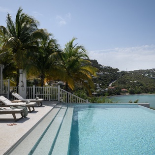 Stunning infinity pool overlooking turquoise bay waters, framed by swaying palms and hillside homes in this tropical paradise setting.