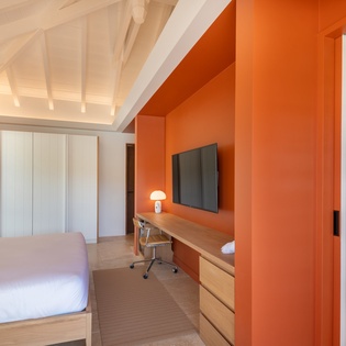 Relax in your stylish bedroom featuring a vibrant orange accent wall, exposed beam ceiling, and modern workspace perfect for unwinding.