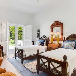 Step into your bright retreat with twin beds dressed in crisp linens and decorative pillows, where French doors open to lush gardens, blending indoor comfort with outdoor beauty.