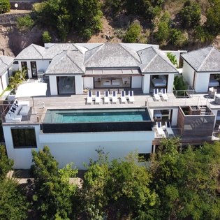 Aerial view of a modern hillside villa featuring contemporary architecture, multiple terraces, and a private infinity pool surrounded by tropical landscaping and natural terrain.