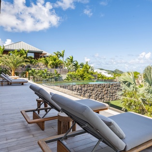 Spacious wooden deck with comfortable loungers overlooking tropical gardens and neighboring villas under bright blue skies.