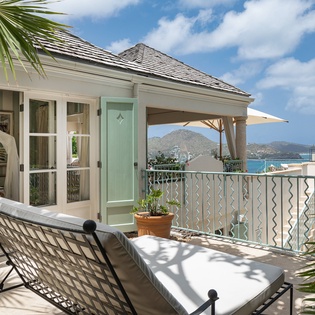Relax on your private terrace with cushioned lounge chairs framed by tropical palms and a soothing ocean breeze, with serene views of the harbor and rolling hills beyond.