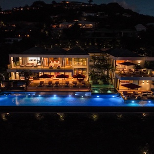 Modern luxury villa with infinity pool illuminated against hillside backdrop at twilight.