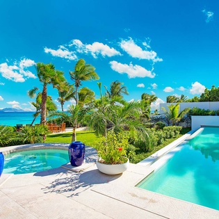 Stunning beachfront property featuring dual pools surrounded by swaying palms and turquoise ocean views, with lush gardens creating a private tropical paradise under endless blue skies.
