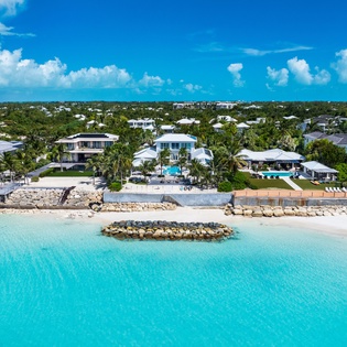 Stunning beachfront properties line pristine turquoise waters, framed by lush tropical vegetation and clear blue skies, creating an idyllic island retreat setting.