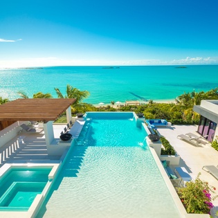 A stunning beachfront villa featuring modern architecture, a pristine swimming pool, and lush tropical gardens overlooking crystal-clear turquoise waters.
