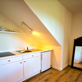 Your private kitchenette tucks neatly under the eaves, featuring a sleek cooktop, sink, and charming shelf-displayed teaware—perfect for morning coffee or light meals in your cozy retreat.