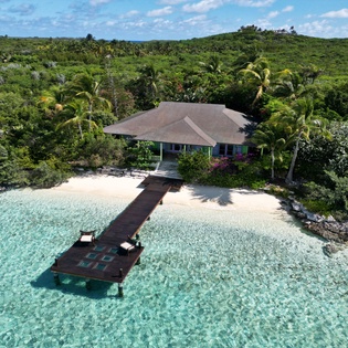 Stunning aerial view of a tropical waterfront villa with private dock extending into crystal-clear turquoise waters, surrounded by lush island vegetation.