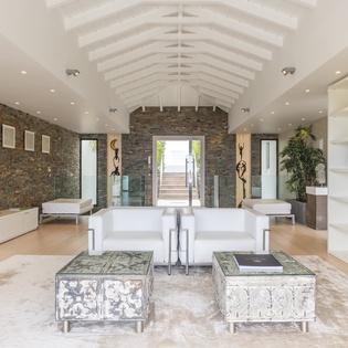A dramatic vaulted ceiling with exposed beams soars above this sophisticated living space. Modern white furnishings create a peaceful retreat against textured stone walls and warm wood flooring.