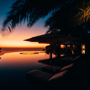 Tropical sunset paints the sky in brilliant oranges and blues over an infinity pool surrounded by swaying palms.