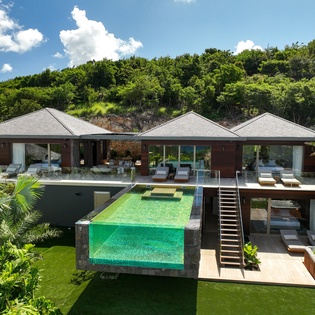 Modern tropical villa featuring dramatic infinity pool with glass walls, surrounded by lush hillside vegetation.