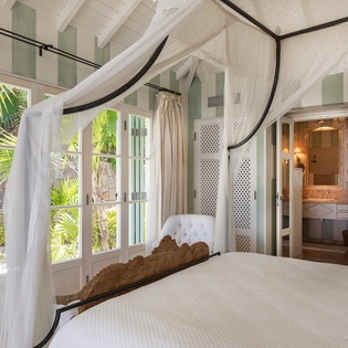Step into your serene tropical retreat, where a romantic canopy bed frames lush garden views and your private ensuite awaits beyond elegant latticed doors.