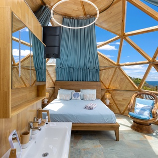 Step into your unique geodesic sanctuary where stunning ocean views surround you through floor-to-ceiling windows. Natural light floods the space, highlighting warm wood details and the inviting bed that awaits your arrival.