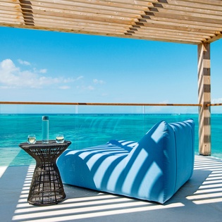 Unwind in your private oceanfront lounge, where striped cushions and wine await under elegant shade with endless turquoise waters stretching before you.