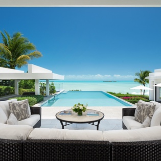 A pristine modern villa featuring an infinity pool overlooking turquoise Caribbean waters. Elegant outdoor seating with plush white cushions invites relaxation under swaying palms in this luxurious beachfront retreat.
