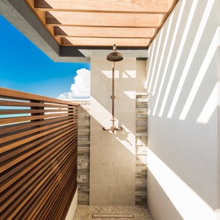 This stunning outdoor shower sits beneath a wooden-slatted pergola, open to clear blue skies. Clean stone walls and warm wood slats create a private, spa-like retreat.