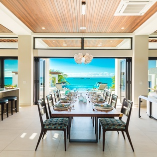 Step into an elegant dining space where the warm wood ceiling frames sweeping ocean views. Open doors invite the sea breeze in, while seating for ten promises unforgettable meals with the turquoise water as your backdrop.