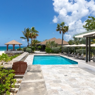A stunning beachfront property featuring a pristine pool, shaded lounge areas, and direct access to turquoise waters and white sand. Tropical palms frame the tranquil setting.