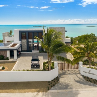 A stunning modern villa perched on turquoise shores, where contemporary architecture meets tropical paradise. Palm trees frame the entrance to this exclusive beachfront retreat.