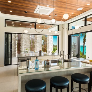 Your culinary haven features a stunning kitchen with a sleek island, modern appliances, and breathtaking turquoise sea views framed by expansive glass doors—perfect for preparing meals while soaking in the beauty outside.