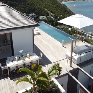 Elevated oceanfront property features a spacious deck with infinity pool, outdoor dining, and modern white-and-wood architecture overlooking a picturesque bay surrounded by lush hillsides.