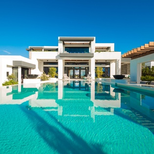 A striking modern villa set in pristine surroundings, featuring a stunning turquoise pool and architectural elegance against a brilliant blue sky.