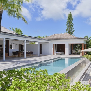 Modern tropical villa with private pool surrounded by lush greenery and swaying palms under a brilliant blue sky.