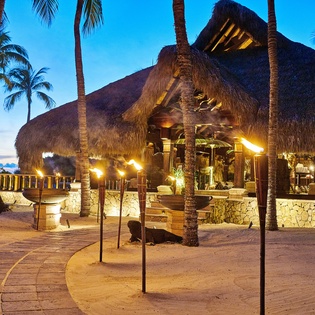 Tropical beachside restaurant with thatched roof and stone walls illuminated at dusk, surrounded by swaying palm trees.