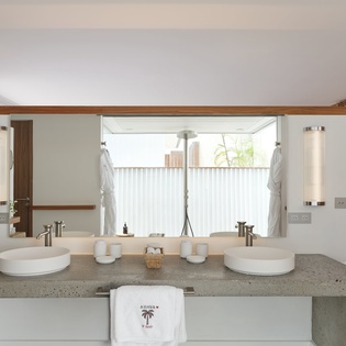 Step into your elegant dual-sink bathroom with polished concrete counters, modern fixtures, and a tranquil window view. Fresh robes await your arrival.