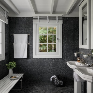 Refresh in your elegant spa-like bathroom featuring dramatic dark tile, double sinks, and tropical garden views through charming windows.