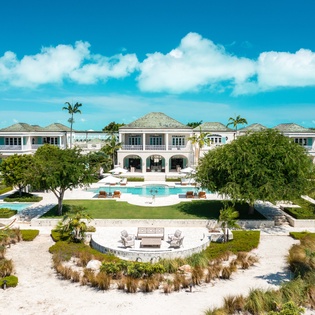 An elegant tropical estate featuring grand colonial architecture, lush manicured gardens, and a sparkling pool set against brilliant blue skies and surrounding greenery.