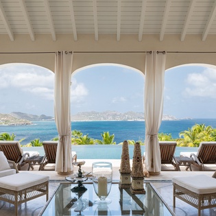 Sink into plush wicker loungers and let your gaze drift across the sparkling turquoise waters and distant hillsides framed by elegant arches—your own private paradise.