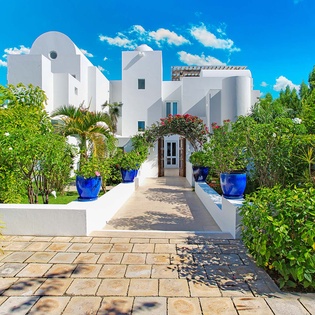 A striking white villa with distinctive architecture sits surrounded by vibrant tropical gardens, blue planters, and flourishing greenery under a brilliant sky.