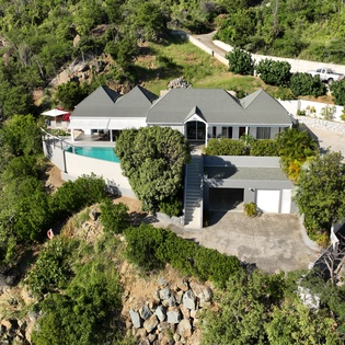 Aerial view of modern vacation villa surrounded by lush tropical vegetation, featuring swimming pool and private parking area.