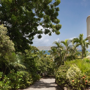 A winding path flanked by lush tropical gardens leads toward tranquil coastal views. The property's white exterior and elegant balcony blend seamlessly with the surrounding greenery.
