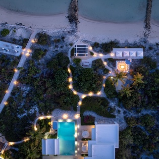 Aerial view of a beachfront property illuminated at dusk, showcasing the swimming pool, pathways, and structures along pristine white sand and turquoise waters.