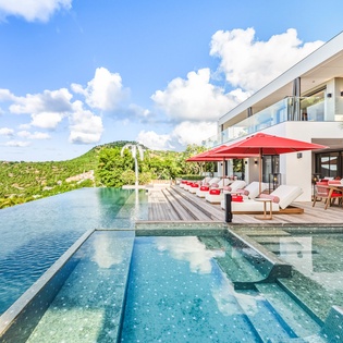 Modern luxury villa with infinity pool overlooking lush tropical hills under bright blue skies.