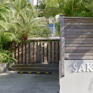 Modern entrance to Sakura property featuring tropical landscaping and contemporary architectural design amid lush palm trees.